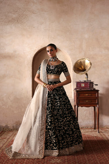 Taseer dark green velvet lehenga choli with gold zari jaal and ivory tissue dupatta, standing by a haveli arch