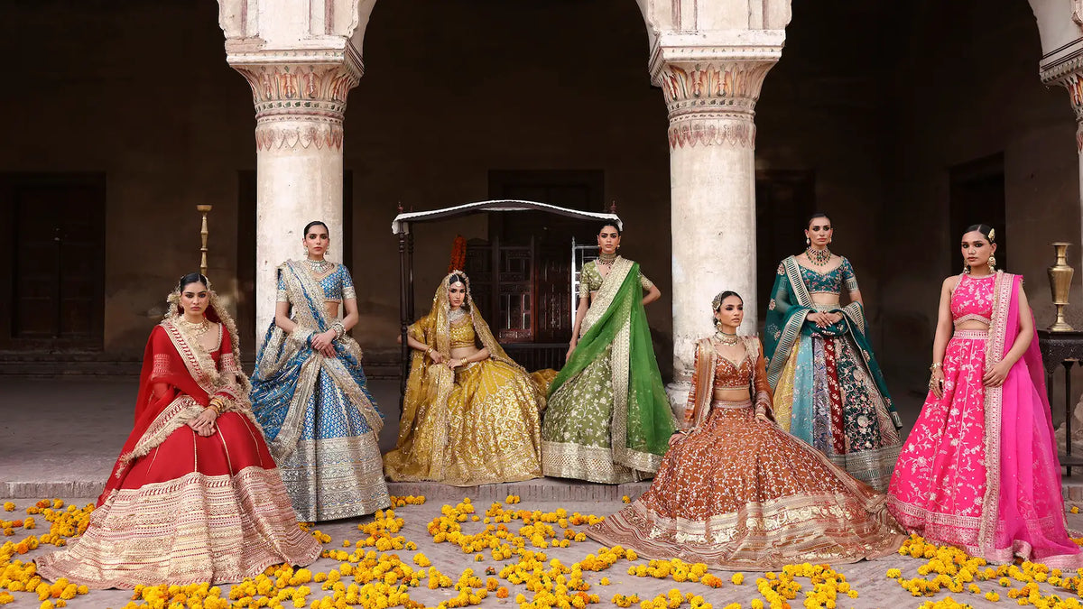 Models in red, gold, green, teal and pink bridal lehenga cholis among marigold petals in a Mughal courtyard, Waqas Shah lehenga choli collection